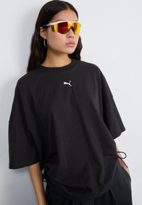 Svart oversized T-skjorte laget av bomull, med en hvit Puma-logo på brystet og snor detaljer i kanten. Brukt med fargerike solbriller.