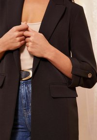 Frau, die die Ärmel eines schwarzen Blazers anpasst, trägt ein beiges Oberteil, hoch taillierte blaue Jeans und einen schwarzen Gürtel mit goldener Schnalle.