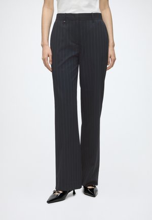 Femme portant un pantalon noir à larges jambes à fines rayures et des talons noirs pointus, debout, les mains détendues le long du corps.