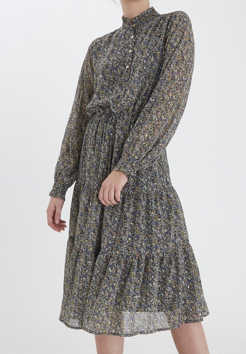 Bloemenjurk met lange mouwen, hoge hals en knoopsluiting. De stof heeft een blauw, groen en geel patroon, met een geplooide rok.