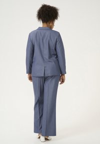Completo azzurro a righe con giacca su misura e pantaloni a gamba larga, realizzato in tessuto leggero con una texture liscia. Scarpe bianche visibili.