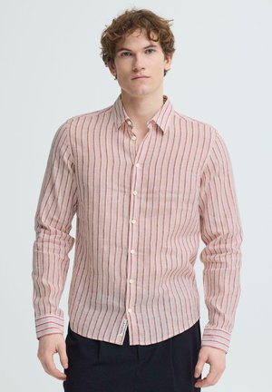 Ung mand med krøllet hår iført en lyserød langærmet skjorte med lodrette røde striber og mørke bukser, stående foran en ensfarvet baggrund.