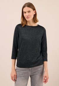 Frau mit schulterlangem, glattem Haar, die ein schwarzes, glitzerndes Langarmoberteil und hellgraue Jeans trägt, vor einem hellen Hintergrund.