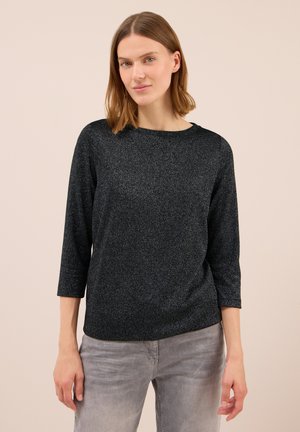 Femme avec des cheveux lisses jusqu'aux épaules portant un haut à manches longues noir pailleté et un jean gris clair sur un fond uni.