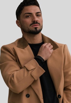 Brown double-breasted coat with a brown collar over a black sweater. Accessories include a black bracelet. Model has short dark hair and trimmed beard.