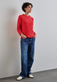 Maglione rosso con texture e scollatura rotonda, abbinato a jeans a gamba larga blu e sneakers bianche. Modella in piedi davanti a uno sfondo neutro.