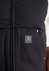 Černá péřová vesta s předním zipem, dvěma velkými kapsami a čtvercovou nášivkou loga Carhartt na levé hrudi. Hladký textilní povrch.