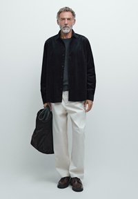 Sort kordfløjl skjorte med knapper, grå sweater, cremefarvede bukser, mørkebrune sko, holder en brun taske. Neutral baggrund, helkropsvisning.