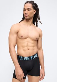 Μυώδης άνδρας με ραβδώσεις, φορώντας μαύρα μποξεράκια Calvin Klein, κοιτάζει προς τα αριστερά του σε ένα απλό ανοιχτό φόντο.