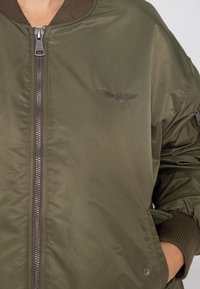 Veste bombardier vert olive à finition brillante, avec col et poignets côtelés, fermeture éclair sur le devant, et un discret logo brodé en forme d'aile sur la poitrine.