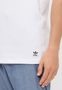 Valge ribaga puuvillane T-särk lühikeste varrukatega ja must silt, millel on Adidas logo alumises vasakus nurgas. Üksikasjalik disain, mugav lõige.