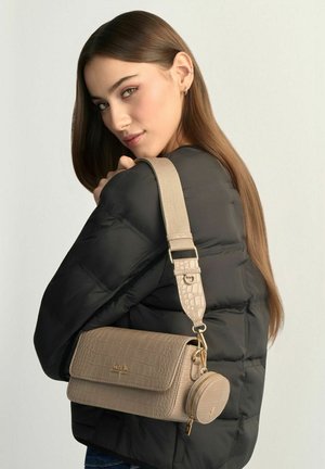 Młoda kobieta w czarnej kurtce puchowej niesie beżową torebkę na ramię o teksturze krokodyla z odpinaną okrągłą sakiewką na monety, spoglądając przez ramię.