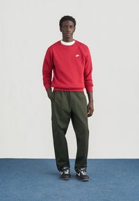 Crvena majica s kapuljačom s bijelim Nike logotipom, uparena s širokim maslinastozelenim cargo hlačama i crnim atletskim tenisicama. Jednostavan, moderan dizajn.