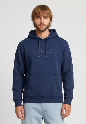 Mężczyzna o jasnobrązowych włosach stoi zwrócony przodem, ubrany w granatową bluzę z kapturem marki Kaporal i jasnoniebieskie dżinsy na jednolitym tle.