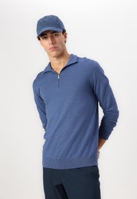 Blue knitted sweater with a quarter-zip collar, ribbed cuffs, and hem. Paired with a blue cap featuring a small logo on the front.