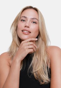 Goldring mit kleinen, klaren Edelsteinen, der an einer Hand getragen wird, die sanft das Kinn berührt. Das Model hat lange, wellige blonde Haare und ein neutrales Make-up.