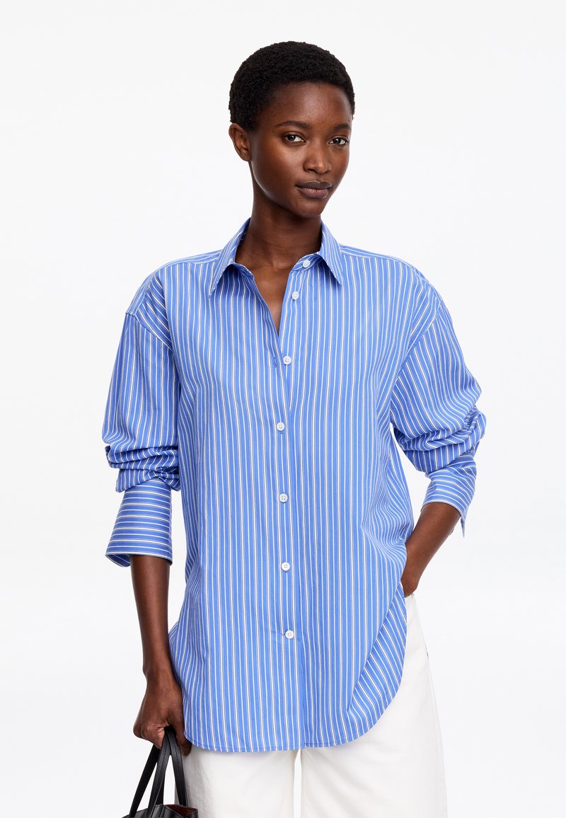 Femme portant une chemise à rayures verticales bleues et blanches avec manches retroussées, pantalon blanc, tenant un sac noir dans sa main gauche.