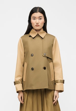 Žena v dvojfarebnom béžovom a hnedom dvojradovom kabáte s gombíkmi a v zodpovedajúcej plisovanej sukni, stojaca na jednofarebnom pozadí.