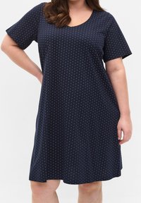 Kobieta w granatowej sukience z krótkim rękawem i okrągłym dekoltem, ozdobionej małymi białymi kropkami, stojąca z jedną ręką na biodrze.