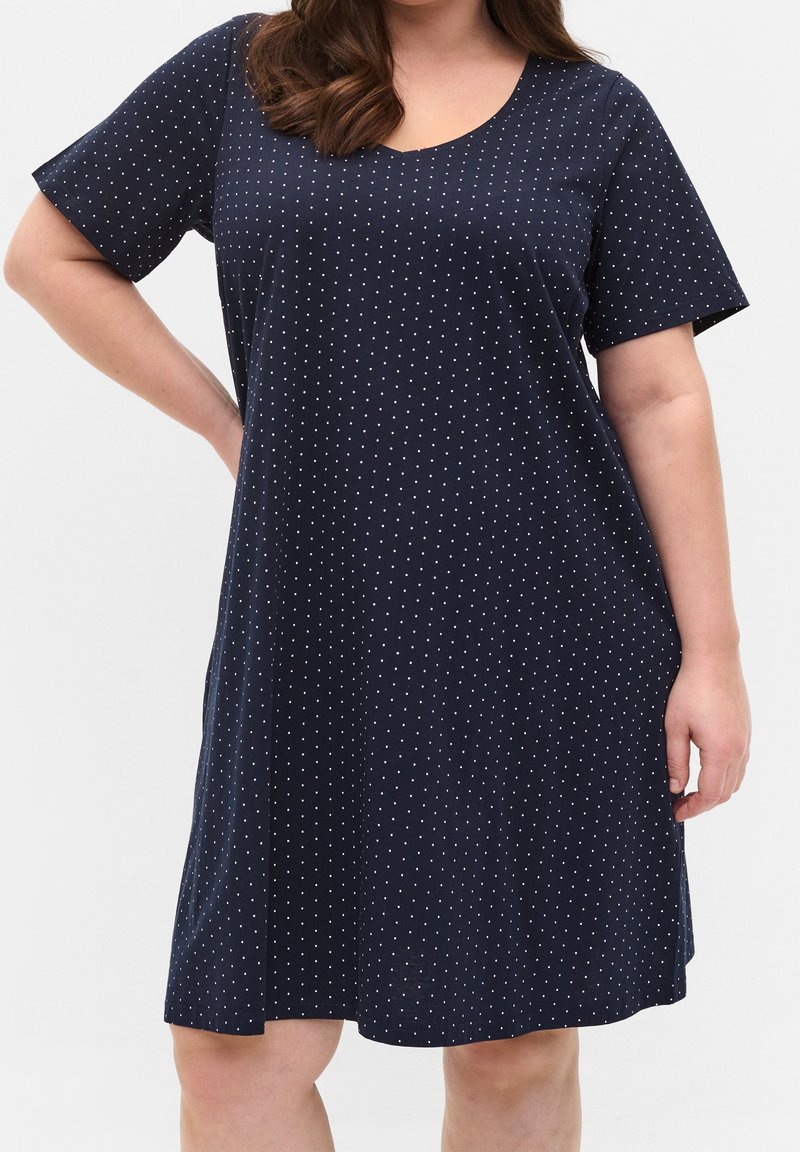 Kobieta w granatowej sukience z krótkim rękawem i okrągłym dekoltem, ozdobionej małymi białymi kropkami, stojąca z jedną ręką na biodrze.