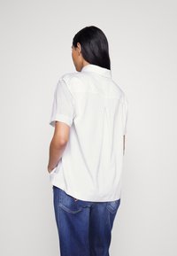 Vit kortärmad skjorta med en rundad fåll, utrustad med en bakåtvikt veck och en klassisk krage. Kombinerad med löst sittande blå jeans.