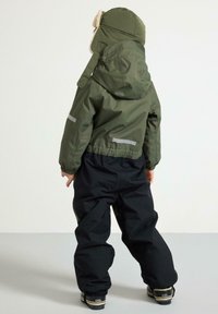 Giacca impermeabile verde con cappuccio e bordo in pelliccia, abbinata a pantaloni neri. Presenta strisce riflettenti e polsini elasticizzati. Calzature standard.