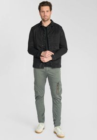 Zwarte spijkerjack, zwart t-shirt, groene cargobroek met ritszakken, witte sneakers met bruine zolen, staand tegen een lichte achtergrond.