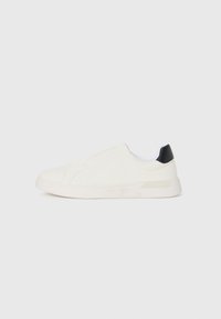 Zapatilla blanca con parte superior de lona lisa, diseño elástico y acento negro en el talón. Cuenta con una suela de goma texturizada.
