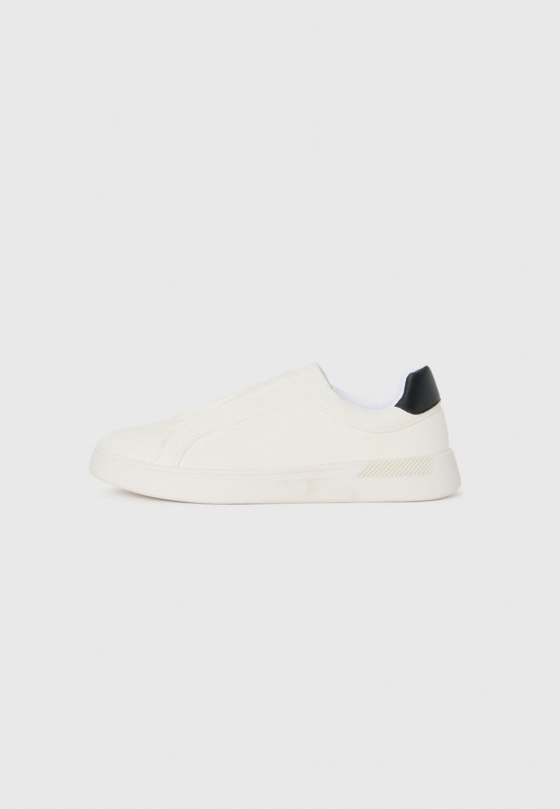 Zapatilla blanca con parte superior de lona lisa, diseño elástico y acento negro en el talón. Cuenta con una suela de goma texturizada.