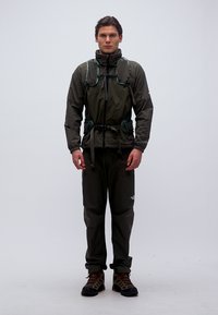 Άνδρας που φοράει σκούρο εξωτερικό μπουφάν, πράσινο γιλέκο με ιμάντες, μαύρο παντελόνι και πεζοπορικά παπούτσια, στέκεται μπροστά σε απλό ανοιχτό φόντο.