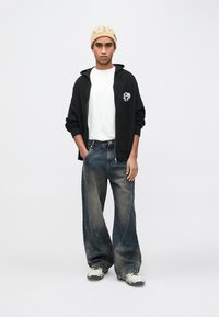 Crni pleteni kardigan na patentni zatvarač preko bijele majice kratkih rukava, uparen s širokim izblijedjelim trapericama i sivim tenisicama. Izgled upotpunjuje bež kapa.