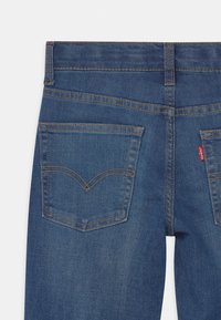 Mörkblå denimjeans med två bakfickor med en krökt sömdesign och en röd etikett på höger ficka. Klassisk femficksmodell.