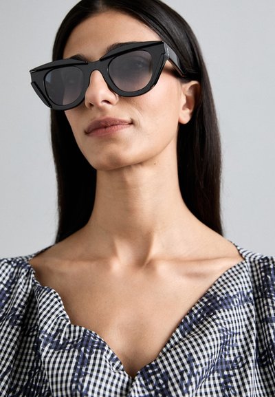Black cat-eye sunglasses with a thick frame and dark lenses. Worn with a patterned gingham blouse featuring a V-neckline.