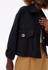 Czarna krótka kurtka z teksturowanej tkaniny z dużymi przednimi kieszeniami i jasnymi guzikami. Zestawiona z musztardową spódnicą.