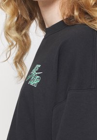 Svart sweatshirt med rundad halsringning. Utrustad med grön text "ONE AIR" och logotypdetalj på vänster bröst. Mjuk, texturerad bomullsvariant.