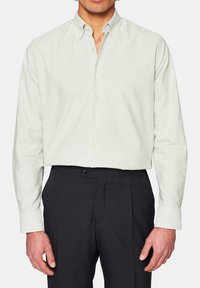 Camicia a maniche lunghe a righe verde chiaro e bianco con colletto a punta, indossata con pantaloni scuri. Presenta una chiusura con bottoni e polsini aderenti.