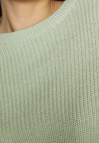 Neulepusero vaaleanvihreänä, leveällä pääntiellä, jossa on teksturoitu ribbineuloskuvio ja pehmeä, kevyt kangas.