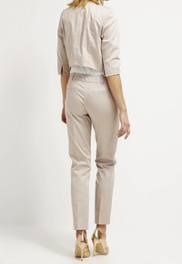 Femme portant un blazer court beige et un pantalon slim assorti, avec des chaussures à talons hauts dorées, se tenant devant un fond blanc uni.