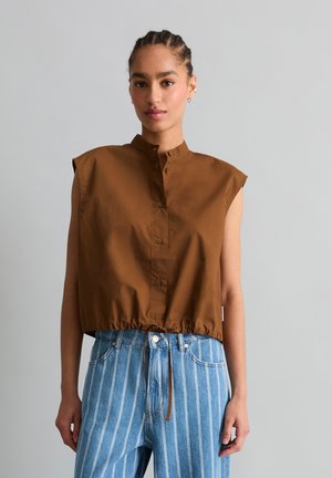 Femme portant un crop top marron sans manches à boutons avec cordon de serrage à l’ourlet et un jean bleu taille haute avec des rayures verticales blanches.