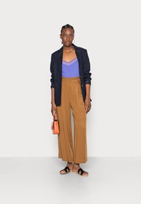 Blazer azul-marinho, blusa lilás clara com detalhe transparente, calças largas castanhas, sandálias pretas de slide, segurando uma pequena bolsa laranja.