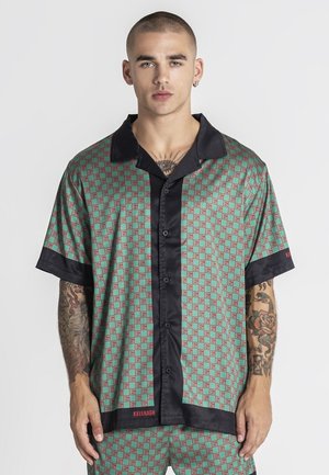 Mladý muž s krátkymi vlasmi a tetovaniami má na sebe zelenú vzorovanú košeľu s krátkym rukávom, čiernym golierom a lemovaním.
