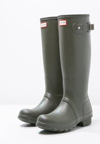 Zelené gumové rain boots s vysokým dizajnom, okrúhlou špičkou, textúrovanou podrážkou a nastaviteľným bočným prackou. Logo "HUNTER" v červenej a bielej farbe.