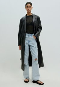Schwarzer Leder-Mantel mit doppelreihigem Design, getragen über einem schwarzen Mesh-Top, kombiniert mit hellblauen zerrissenen Jeans und schwarzen Flip-Flops.