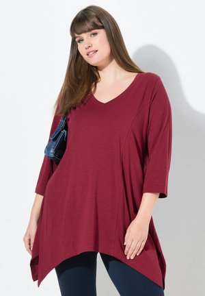 Bordeauxfarbige Tunika mit V-Ausschnitt, Dreiviertelärmeln und asymmetrischem Saum. Leichter Stoff mit glatter Textur. Kombiniert mit dunklen Leggings.