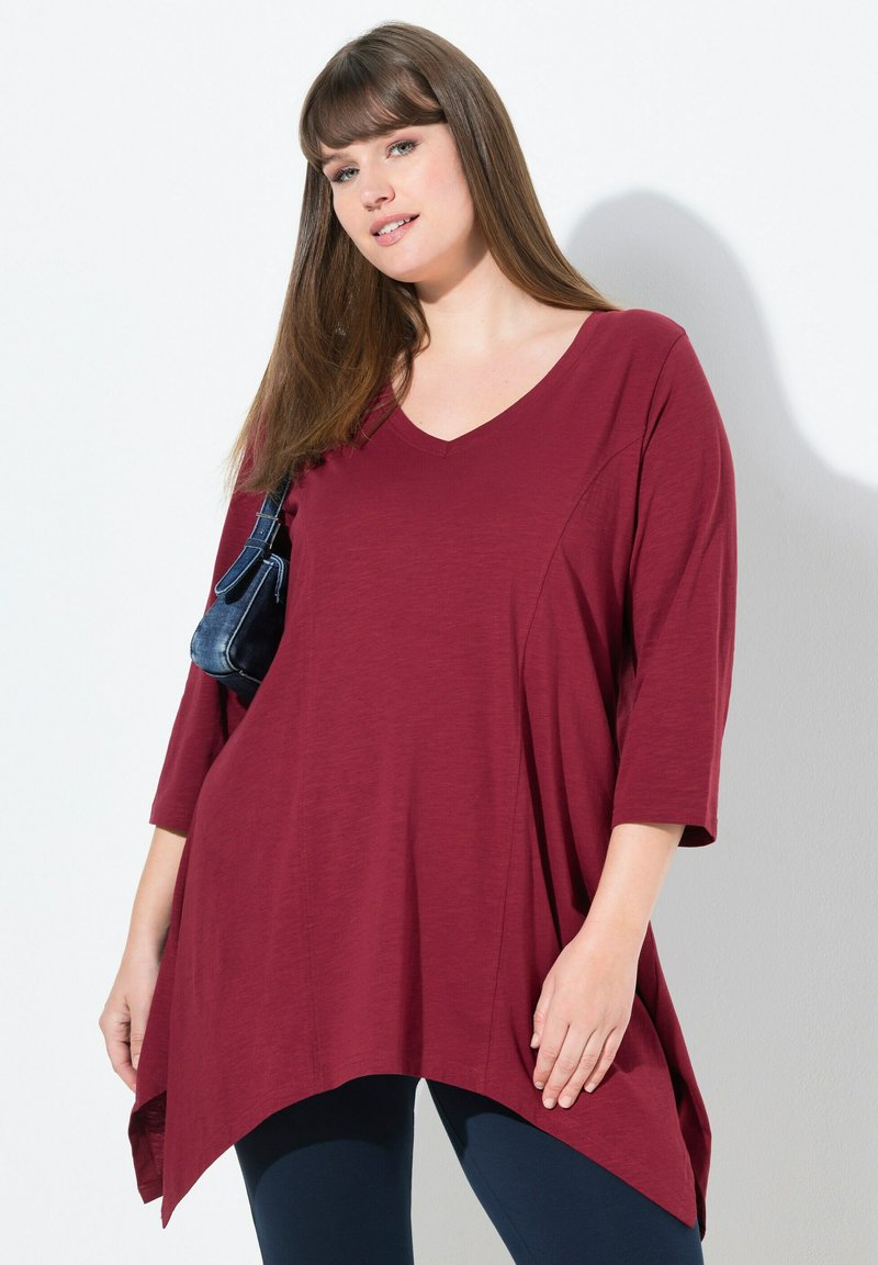 Bordeauxfarbige Tunika mit V-Ausschnitt, Dreiviertelärmeln und asymmetrischem Saum. Leichter Stoff mit glatter Textur. Kombiniert mit dunklen Leggings.