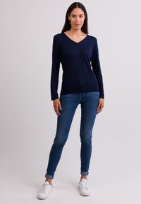 Marinblå långärmad V-ringad tröja i mjukt material med en figurnära design. Återfinns tillsammans med blå skinny jeans och vita sneakers, stående mot en neutral bakgrund.