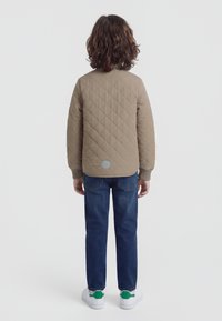 Dziecko z kręconymi brązowymi włosami ubrane w beżową pikowaną kurtkę, niebieskie dżinsy i białe trampki z zielonymi akcentami, stojące tyłem.