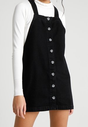 Black button-front pinafore dress worn over a white long-sleeve ribbed top, shown from neck to mid-thigh on a standing person.