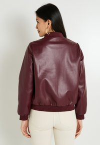Mujer con cabello largo y oscuro, usando pendientes de aro dorados, una chaqueta bomber burdeos brillante y pantalones beige claros, de espaldas a la cámara.