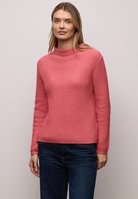 Pull côtelé rose avec un col montant et des manches longues. Associé à un jean bleu foncé, caractérisé par une coupe décontractée et une texture douce.